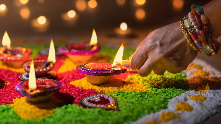 An Indian woman's hand as she lights candles, surrounded by pink, yellow and green petals arranged in a pattern.