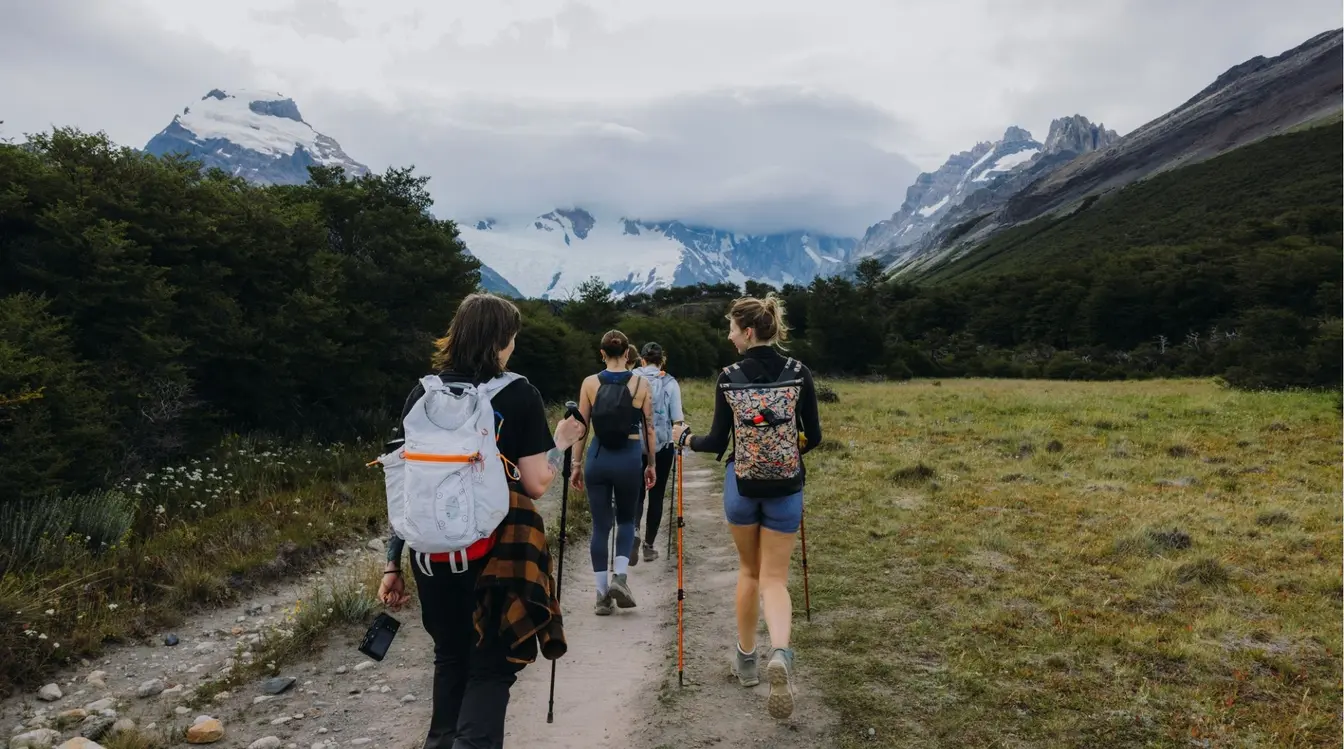 Três pessoas com mochilas caminhando de costas por uma trilha de terra em meio à natureza, com grandes montanhas de picos nevados ao fundo sob um céu nublado.