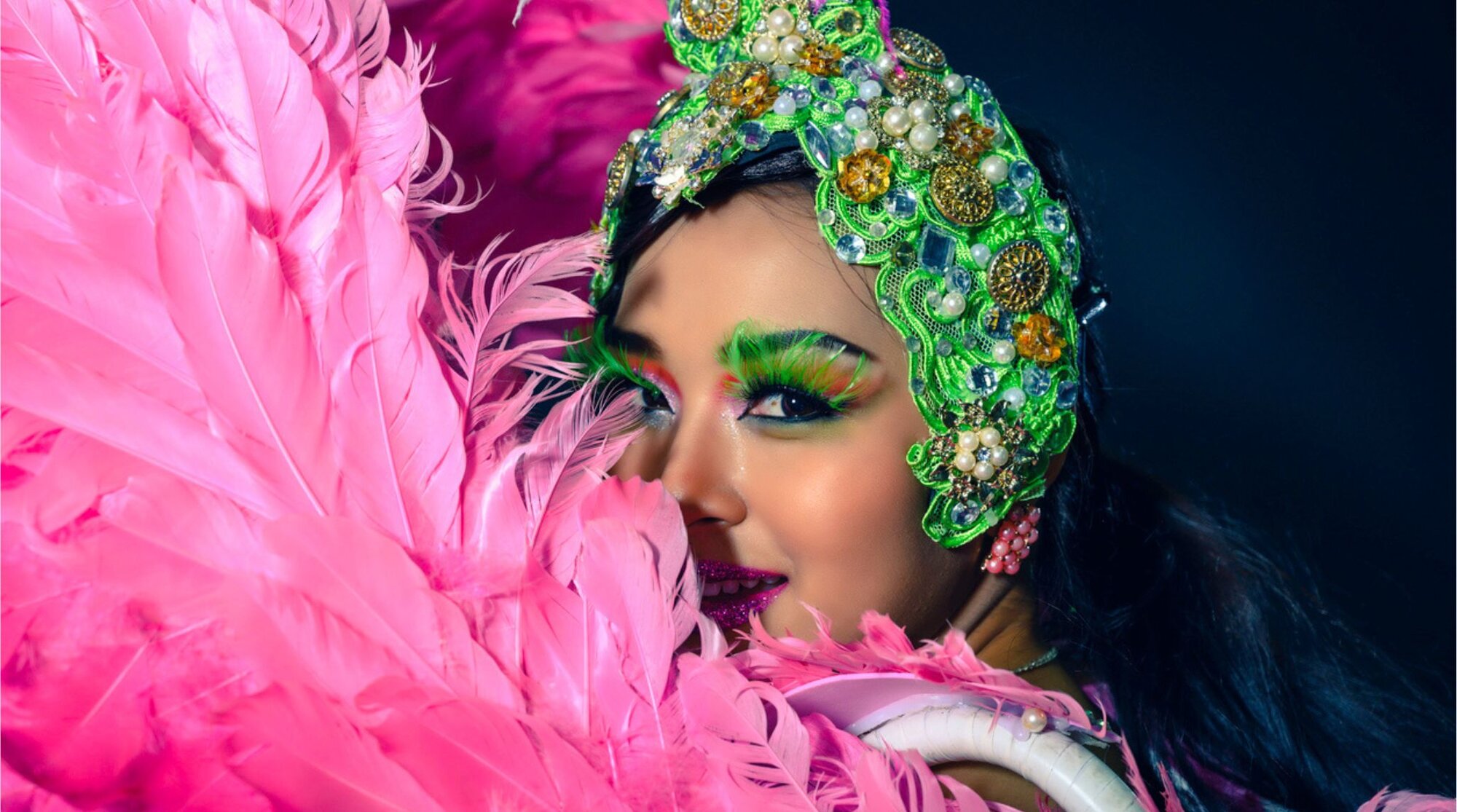 Primer plano de una bailarina tipo showgirl de Las Vegas con tocado brillante y plumas rosas, maquillaje escénico y fondo oscuro