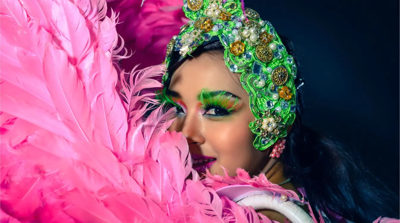 Primer plano de una bailarina tipo showgirl de Las Vegas con tocado brillante y plumas rosas, maquillaje escénico y fondo oscuro