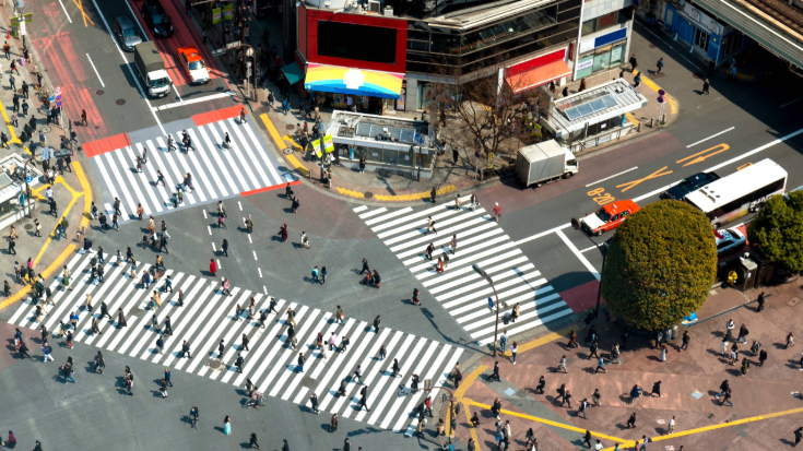 Aerial view of Shibuya Crossing in Tokyo with hundreds of people moving in all directions across wide white-striped crosswalks, surrounded by neon billboards and busy streets — a famous Tokyo Game Show photo spot near the city’s buzzing nightlife.