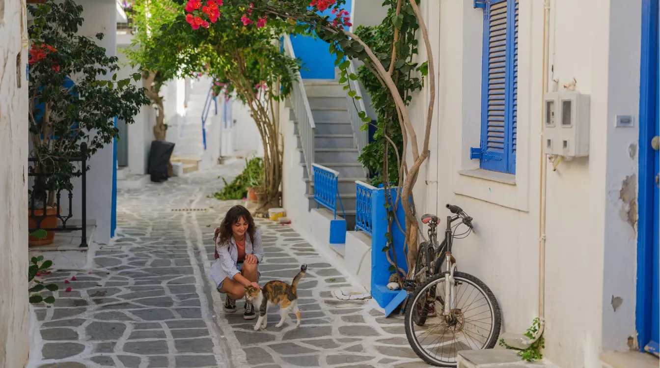 Mulher acariciando um gato nas ruelas de casas brancas e janelas azuis de Mykonos Town, na ilha de Mykonos, Grécia.
