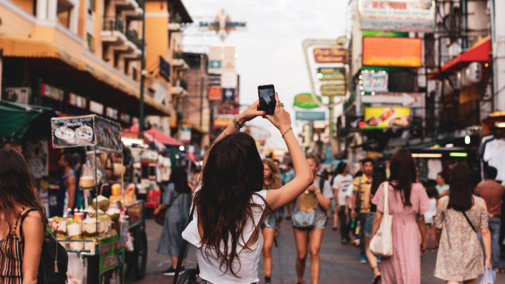 An image of the back of a woman on Khao San Road in Bankgkok, Thailand. She is wearing a white shirt and trying to take a photo of the busy market street and street food vendors in front of her. This is to illustrate a blog post entitled '33 fun facts about Thailand'.