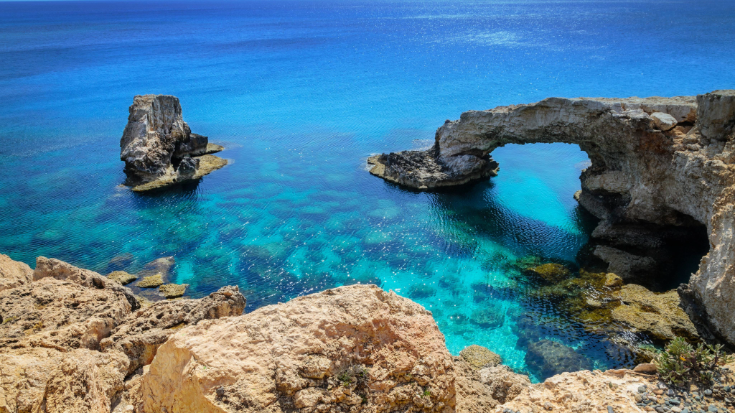 A photograph of a rock arch near of Ayia Napa, Cavo Greco and Protaras on Cyprus - Natural rock arch stretching into the bright blue sea in Cyprus. The water beneath glows with shades of turquoise and emerald, while jagged cliffs frame the scene — a breathtaking spot for cheap hot holidays in October and easy to explore with the best eSIMs for Europe.