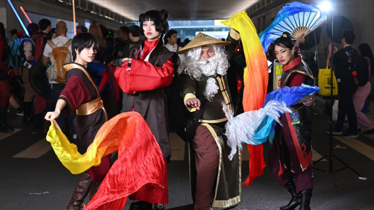 A photograph of four young people, two men and two women, of Asian ethnicity, dressed in cosplay outfits at New York Comic Con. They are dressed in red, orange and black coordinating outfits. One is wearing a triangular straw hat and silver long beard, while one of the women is holding a large sky-blue fan decorated with feathers.