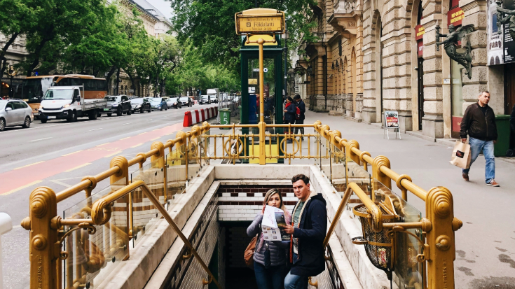 A photograph of the Foldalatti metro stop in Budapest. There is a sign with the metro stop's name on it in the background. In the foreground, there are some stairs leading down into the metro station. On the stairs, a man and woman have stopped and are looking at a map of Budapest. To illustrate a blog post entitled 'The Ultimate Sziget Festival Guide.'