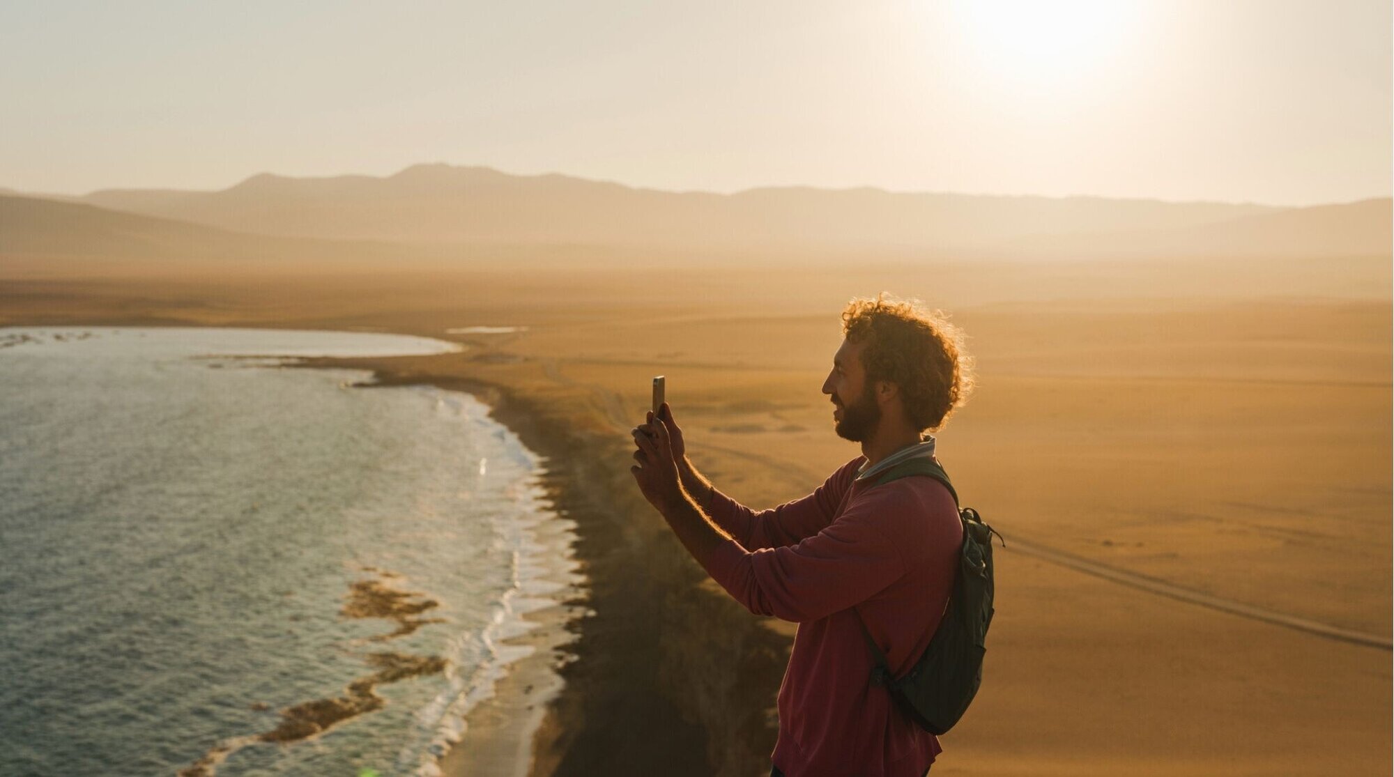 Un viajero fotografía el océano al atardecer desde un mirador junto a una costa amplia y luminosa.