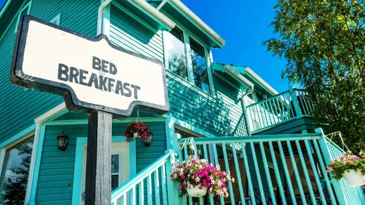 A teal wooden house featuring a rustic white "Bed and Breakfast" sign and hanging flower baskets, symbolising independent, locally-owned accommodation.