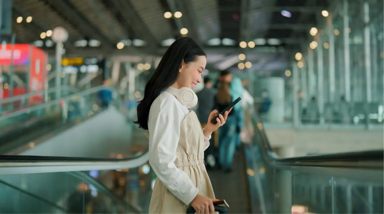 Jovem viajante sorrindo ao usar o celular em uma esteira rolante de aeroporto, com fones de ouvido no pescoço e mala de mão ao lado.