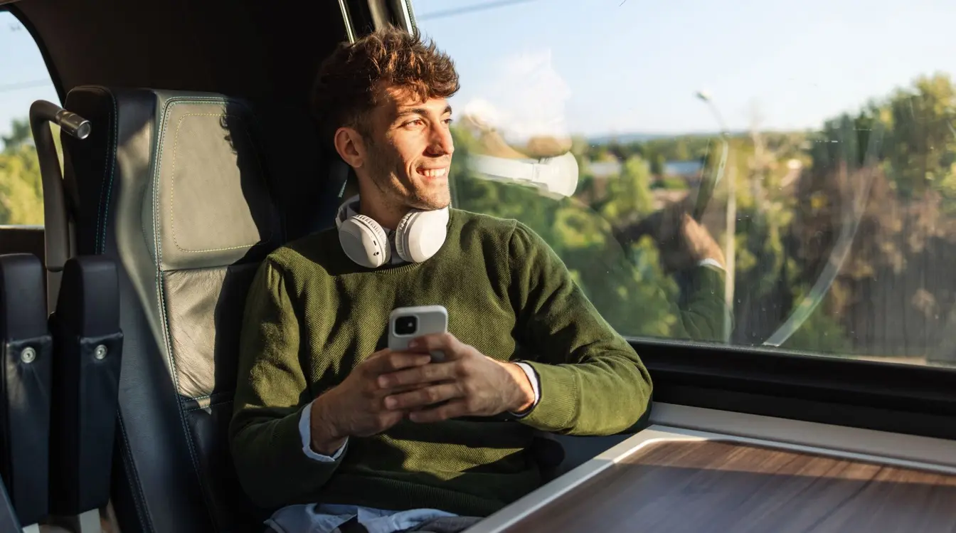 A traveller with headphones around his neck looking out the window of a train.