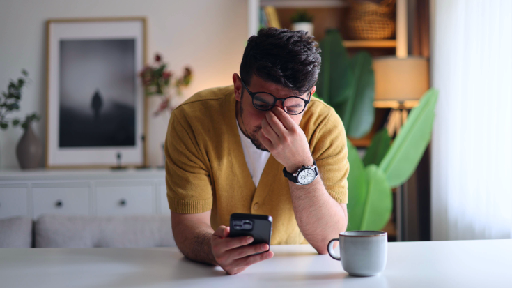 A man leaning on a kitchen counter rubbing his eyes as he looks at his phone screen, to illustrate a blog post entitled 'How to Know if Someone Blocked You on WhatsApp.'