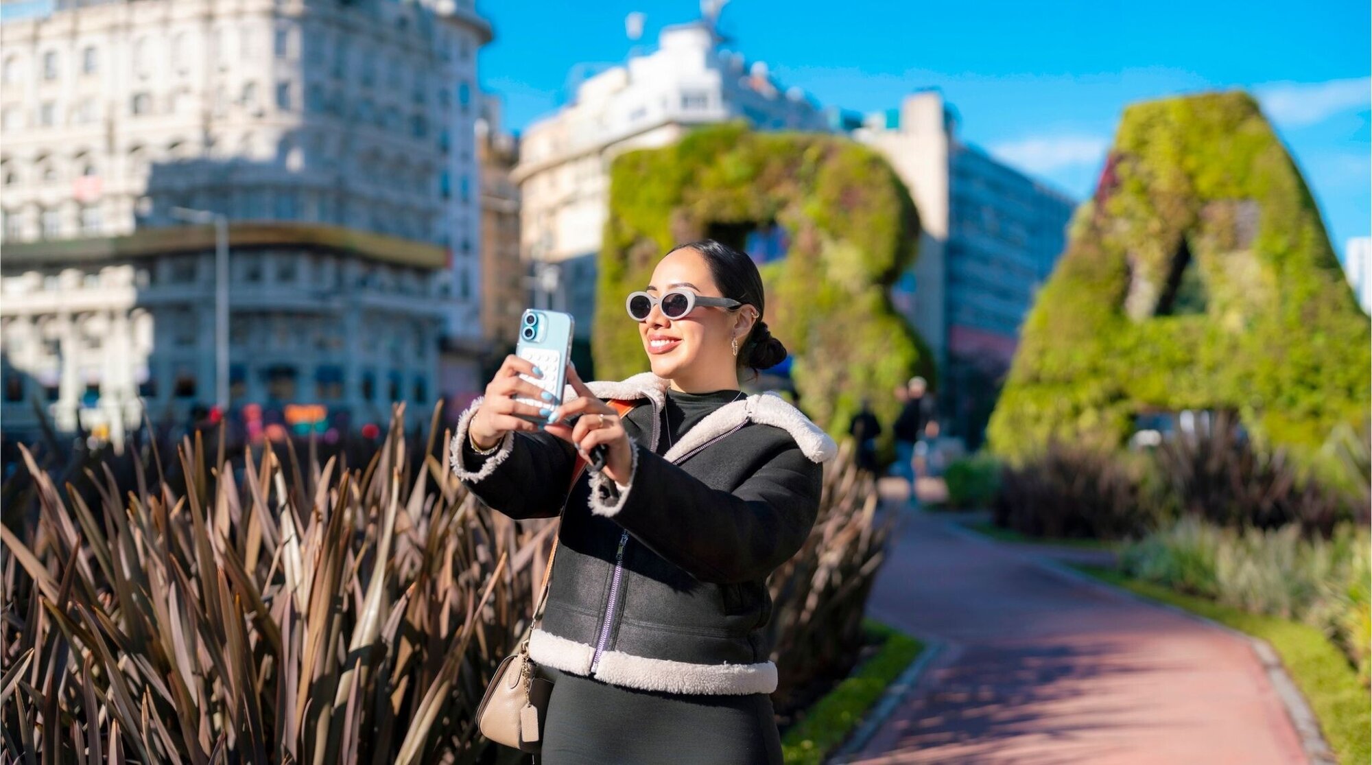 Jovem latina sorridente tirando selfie em Buenos Aires perto do sinal BA e Obelisco em um dia ensolarado, vestindo jaqueta preta na moda e óculos de sol.