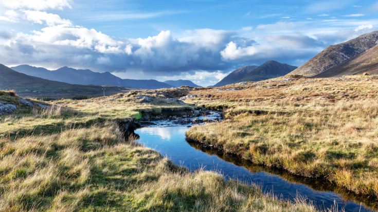 The valleys of Ireland's beautiful and quiet Maumturk Mountains, with a waterway passing through long grass fields.