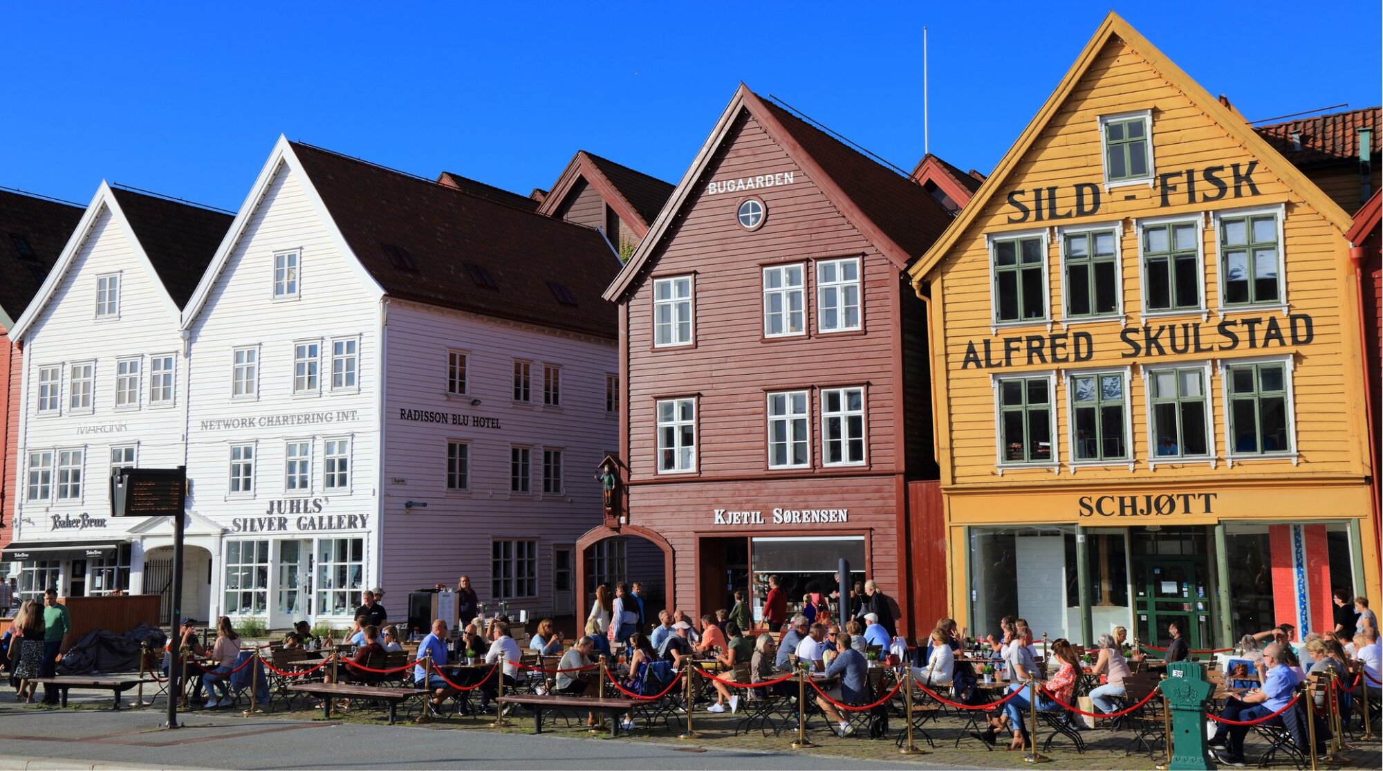 Turistas sentados em mesas ao ar livre em frente às casas de madeira coloridas do bairro de Bryggen, Patrimônio Mundial da UNESCO, em Bergen, Noruega.