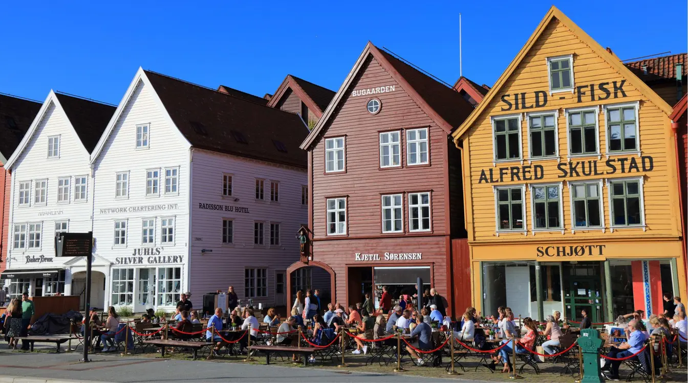 Turistas sentados em mesas ao ar livre em frente às casas de madeira coloridas do bairro de Bryggen, Patrimônio Mundial da UNESCO, em Bergen, Noruega.