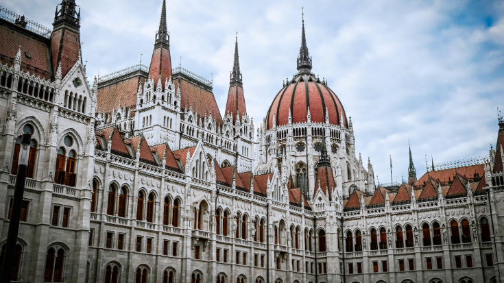 A photograph of Budapest's large Gothic-style parliament building. It shows many windows and the decorative arches above them. The building is in a grey stone colour, while there are domes above the windows in a striking rust/red colour, all against a blue sky with white clouds. To illustrate a blog post entitled 'The Ultimate Sziget Festival Guide.'