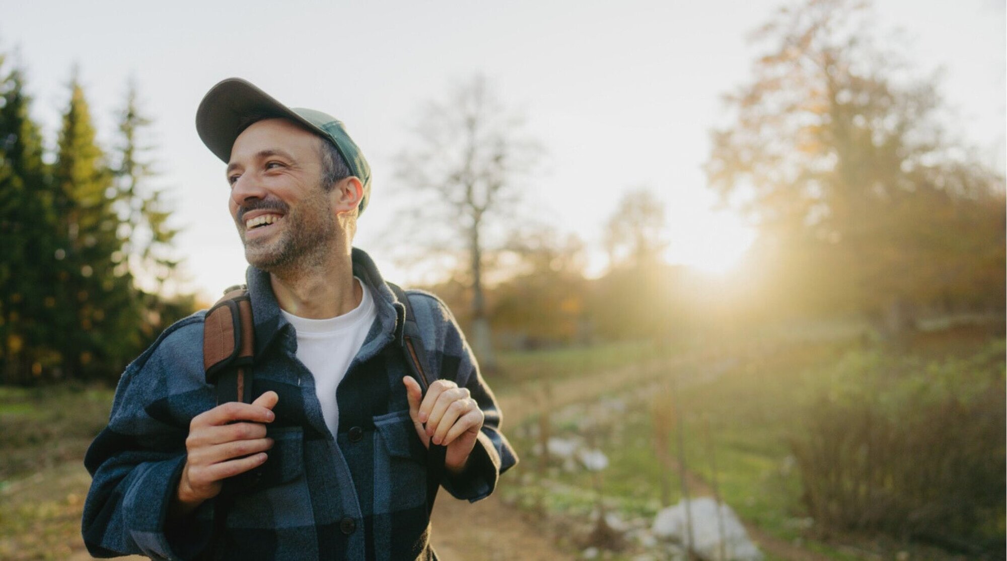 Excursionista sonriendo con mochila en un sendero al aire libre, luz de tarde y paisaje rural.