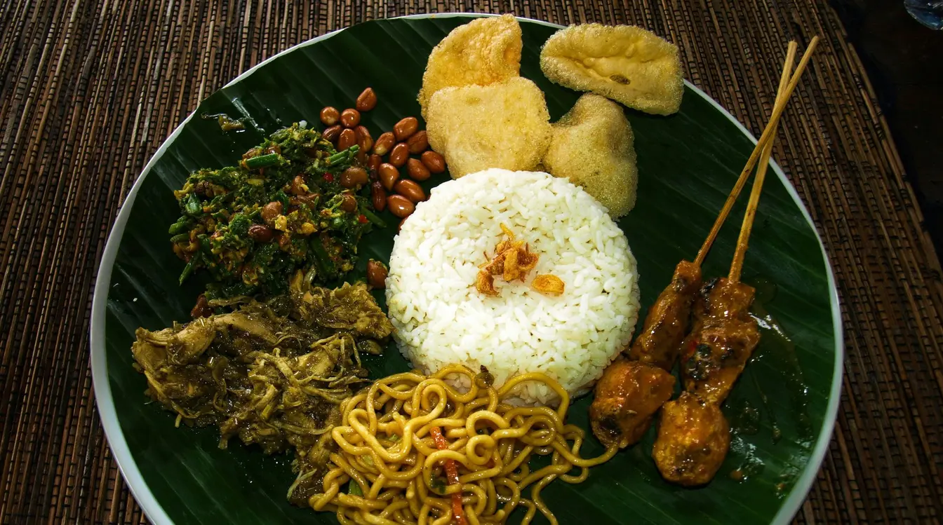 Traditional Indonesian Nasi Campur served on a banana leaf with rice, satay skewers, noodles, peanuts, and other sides.