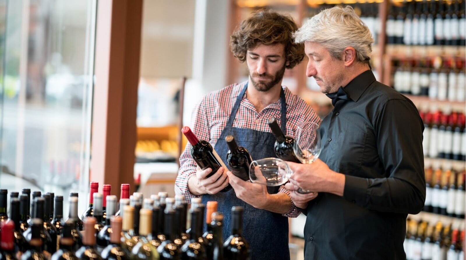 O que comprar na Argentina: Jovem balconista e proprietário da empresa olhando para garrafas de vinho enquanto proprietário detém taças de vinho em uma adega.