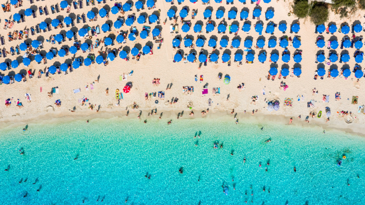 An aerial photograph of a sandy beach in Ayia Napa on the island of Cyprus. The waters are still and crystal clear and the beach is studded with bright blue parasols, with people relaxing underneath and around them. To illustrate a blog post entitled 'where is hot in Europe is October?'