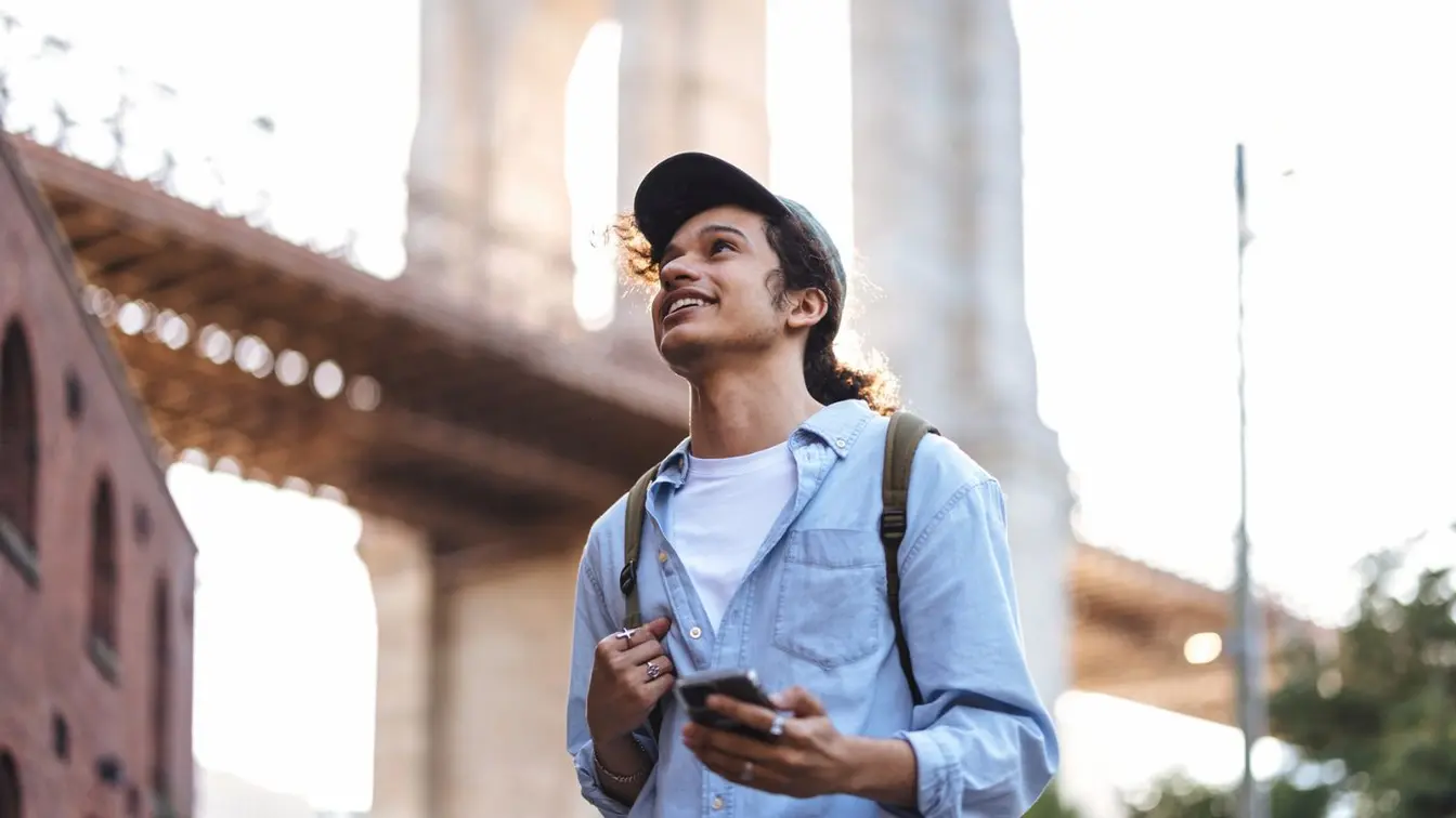 Junger Tourist unter der Brooklyn Bridge lächelt, während er New York City mit Rucksack und Handy mit SIM-Karte für die USA erkundet.