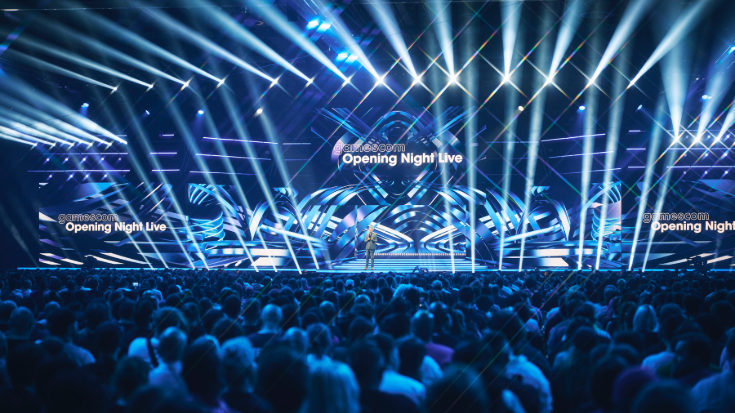 The main gamescom stage from afar, lit with blue laser beams and bright lights, while the packed-out crowd looks on.