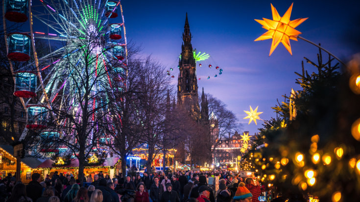 A coloured photograph showing a dark night sky and crowds of people visiting Christmas stalls and amusement rides in Princes Street Gardens in Edinburgh, Scotland. Crowds navigate the busy stalls near the Scott Monument and Ferris wheel in Edinburgh, a popular choice for extreme day trips. To illustrate a blog post entiotled 'Extreme Day Trips - UK Christmas Markets Edition'.