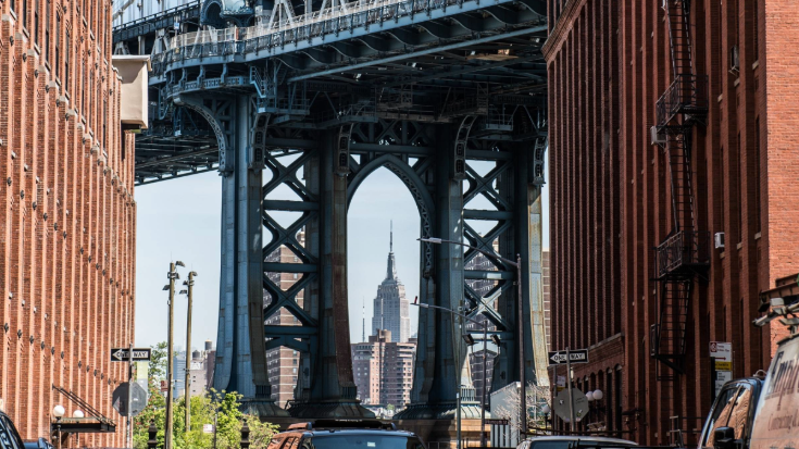 A colour photograph of a famous DUMBO district image, featuring the Empire State Building framed by the Manhattan Bridge. It is a classic New York City view taken from Washington Street in the Dumbo area of Brooklyn. It shows redbrick buildings on either side in the foreground and in the background, the silhouette of the art deco Empire State Building, which is framed by the steel cables and latticework of the Manhattan Bridge. To illustrate a blog post entitled 'New York Comic Con 2025: A Complete Travel Guide for Attendees '.