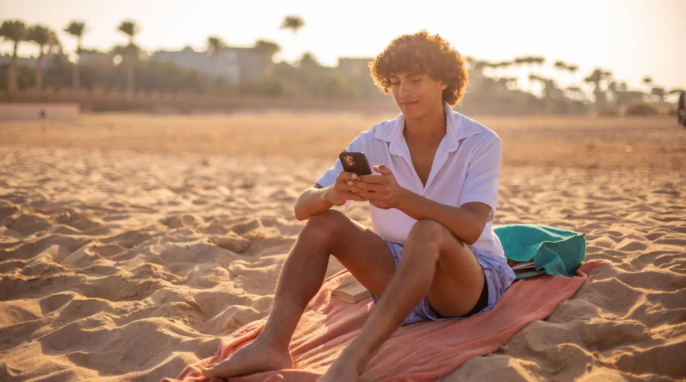 Junger Mann mit eSIM benutzt Smartphone am Strand bei Sonnenuntergang in Ägypten