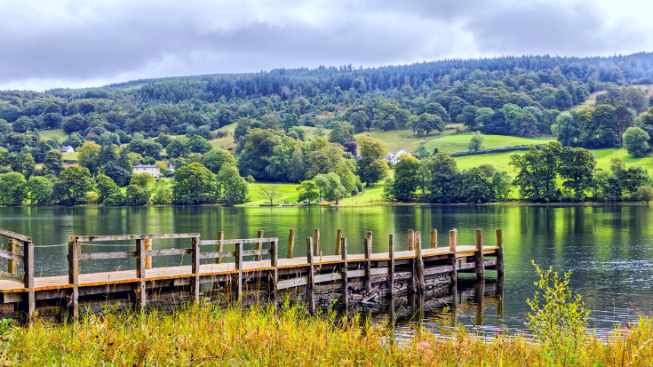 The still waters of Coniston Water in England's Lake District, with green hills in the background and a small brown wooden jetty in the foreground.