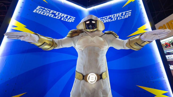 An image of a cosplayer in an all-white trooper style uniform with his face concealed by a black mask for promotion at the Esports World Cup 2024 in Riyadh, Saudi Arabia. Behind him is a blue sign that says 'Esports World Cup'.