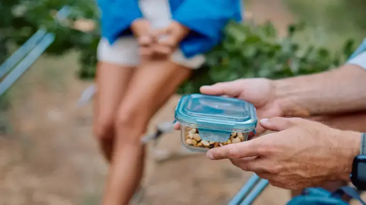 A colour image showing a close-up of a traveller's hands holding a reusable glass container filled with nuts, highlighting zero-waste packing essentials for plastic-free snacking.