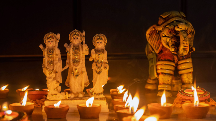 A small stone statue of Lord Rama with diya candles for Diwali next to it, against a dark background.
