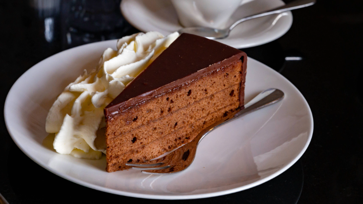 A photograph of a white plate against a black background. On the plate is a traditional Austrian cake called a sachertorte, which is served with coffee in Vienna's iconic coffee houses. The cake is has three slim layers of chocolate sponge, separated by a thin layer of chocolate crea,. the outisde of the cake slice is coated ina. shiny dark brown chocolate ganache and is topped with whipped cream. for a blog post entitled 'Which Country Has the Best Coffee Culture?' 