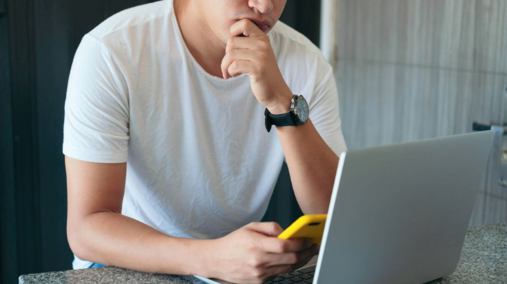 A photograph of a man from sitting at a table and holding a phone with a yellow case, while also sitting in front of a laptop screen. He seems to be deep in thought, is wearing a white t-shirt and a large black and silver watch.This is an image to illustrate a blog post entitled 'Unlimited eSIM: Plans, Benefits, and How It Works'. 