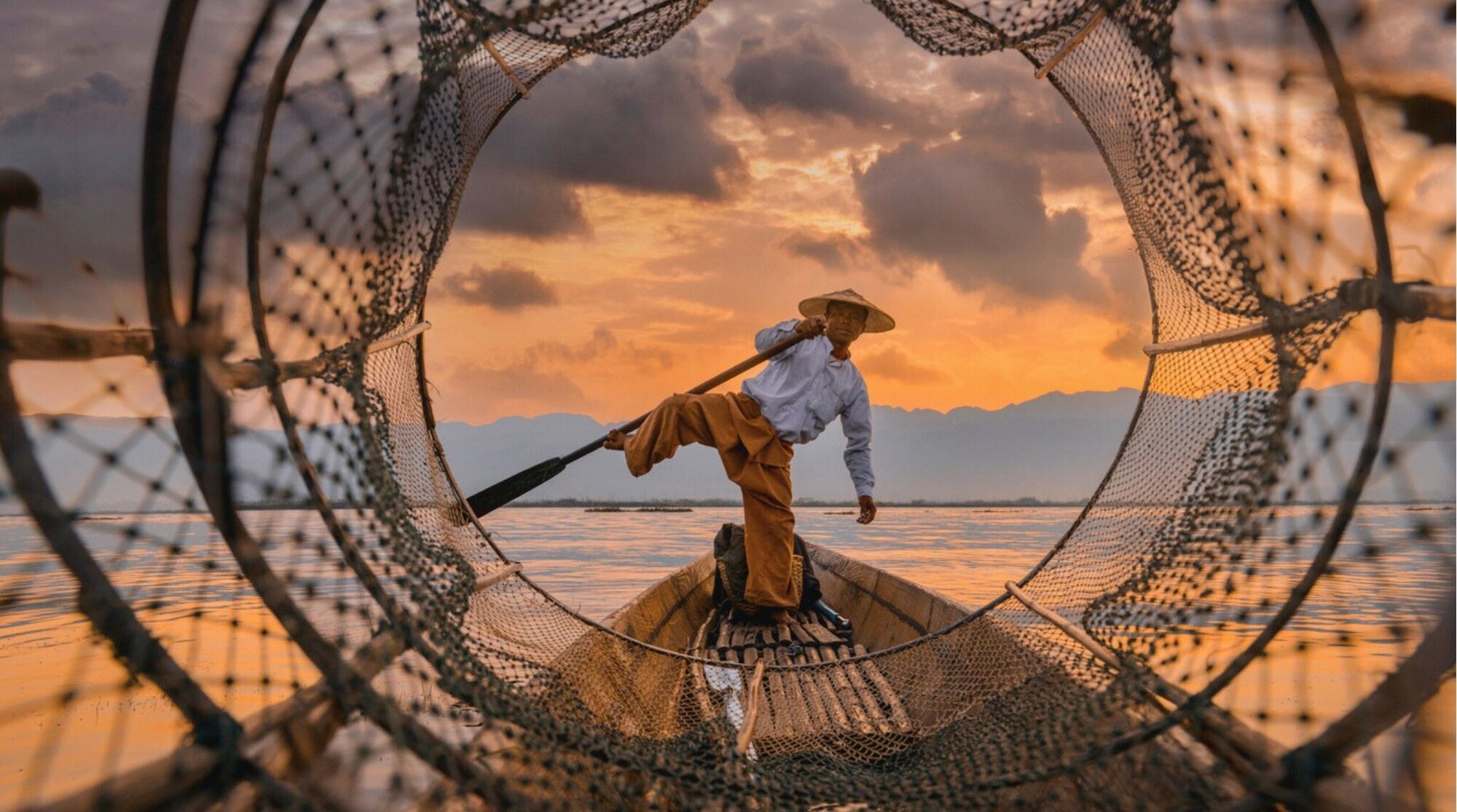 Pescador equilibrándose en una barca al atardecer, red de pesca en primer plano y cielo dorado.
