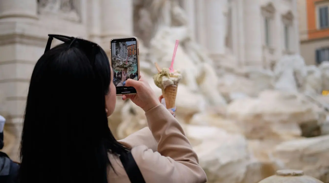 Tourist taking a picture of gelato with her phone in front of Rome’s Trevi Fountain.