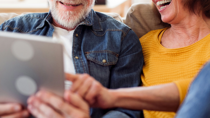 A photograph showing a close-up of a couples' faces as they use an electronic device together, likely a tablet. They are both smiling. He is holding the tablet while she points to something on the screen. He is wearing a blue denim short and has a grey beard and she is wearing a yellow shirt and has bobbed brown hair. This is an image to illustrate a blog post entitled 'Unlimited eSIM: Plans, Benefits, and How It Works'. 