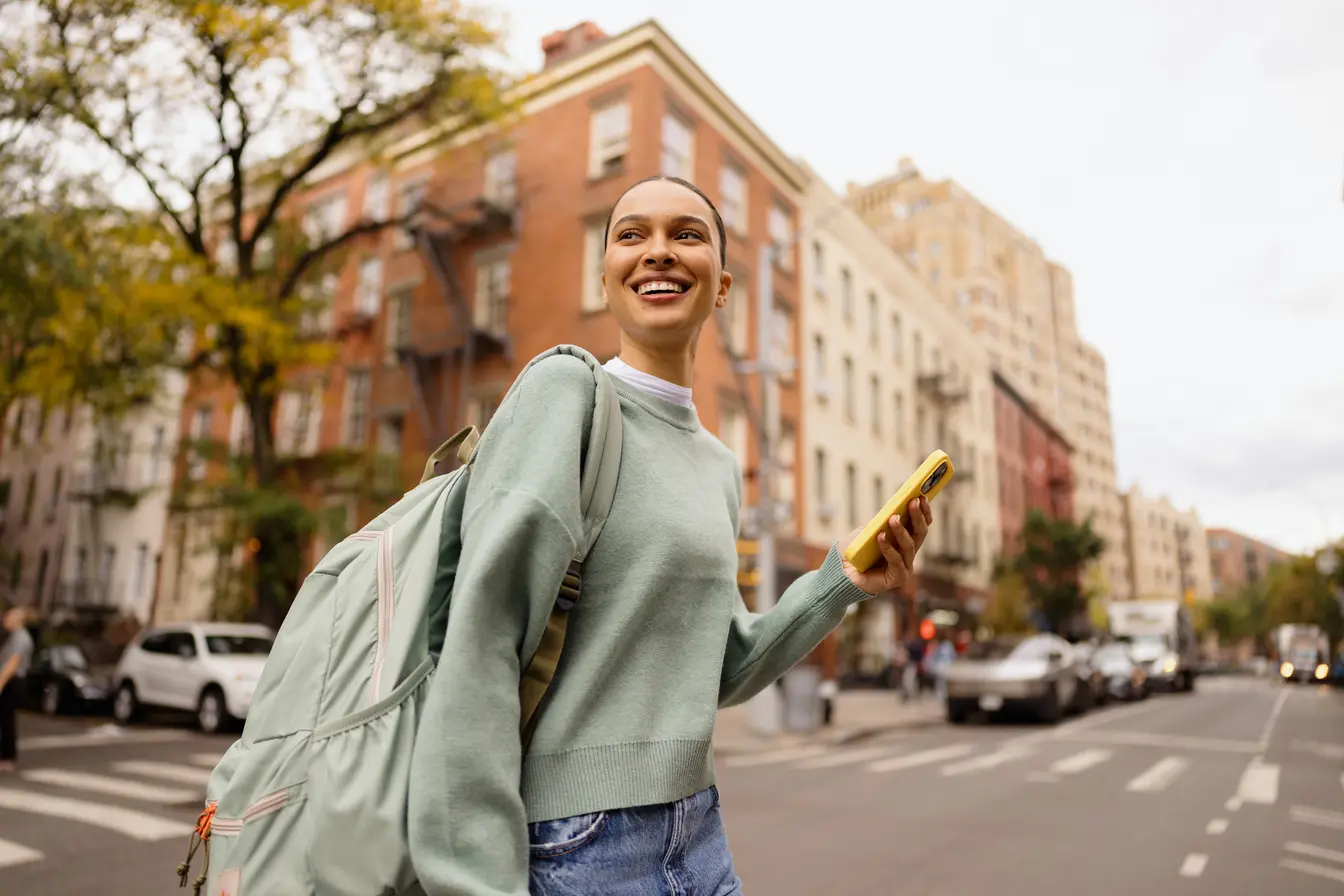 Junge Frau, die auf einer belebten Großstadtstraße entlanggeht, lächelt und hält ein Handy mit einer SIM-Karte für die USA in der Hand.