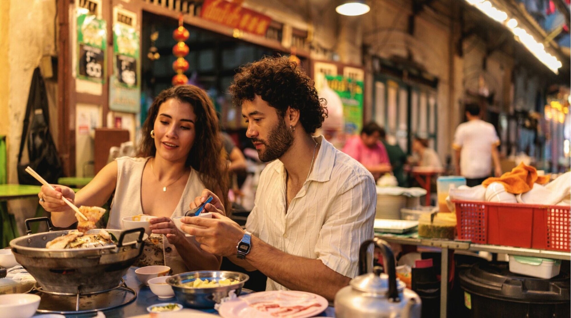Sentados próximos, um casal mergulha ingredientes em uma panela quente e fumegante, aproveitando o calor e os sabores da refeição juntos. Chinatown, em Bangkok, Tailândia.
