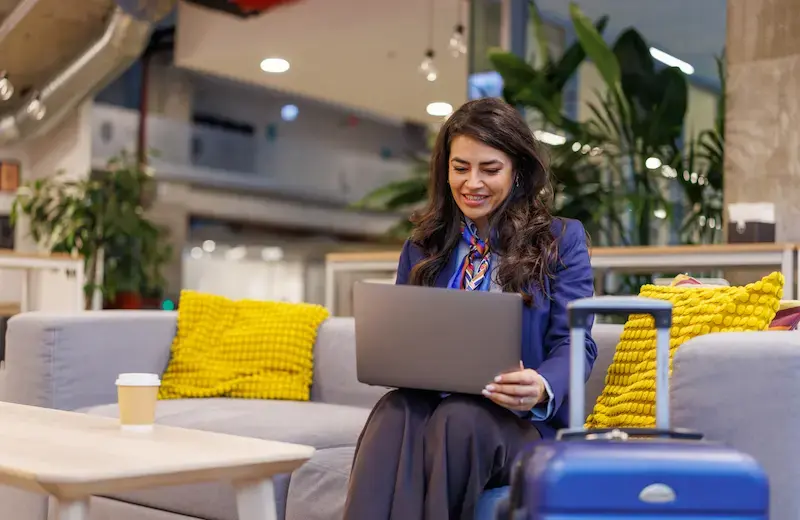 Voyageuse assise dans un espace lounge d’aéroport, travaillant sur un ordinateur portable avec une valise cabine à côté