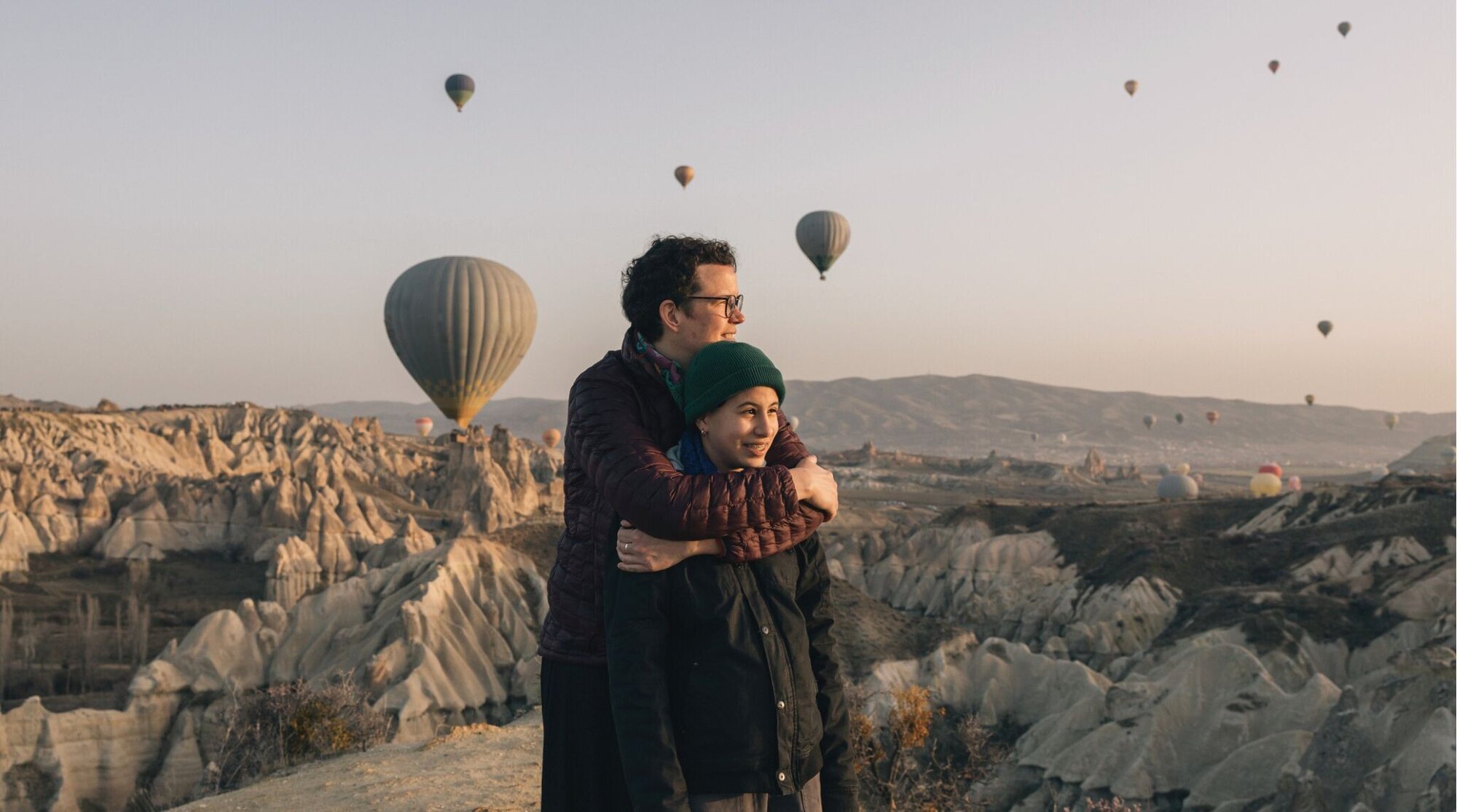 Capadócia. Mulher abraçando sua filha adolescente enquanto observam balões de ar quente voando sobre Göreme