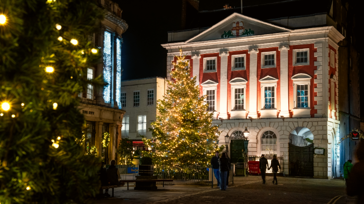 A colour photograph of the historic city of York, England. It's dark and in the foreground are Christmas trees lit up with yellow lights. In the background you can see the red and white painted building York Mansion House, with its columns and sash windows. The scene is located in St Helen's square, York. To illustrate a blog post entitled 'Extreme Day Trips - UK Christmas Markets Edition'.