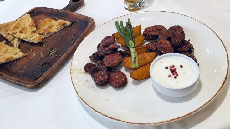 A photograph of some traditional Albanian food, including sliced spicy sausages, flatbreads with rosemary and Albanian beer, all on plates against the backdrop of a tablecloth, to illustrate a blog post entitled Extreme Day Trips from London to Europe.