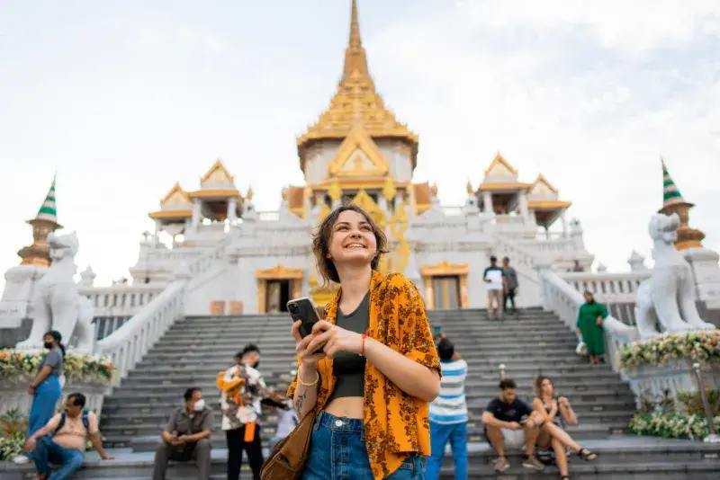 Mujer de pie con un celular en la mano frente a un templo, rodeada de turistas.