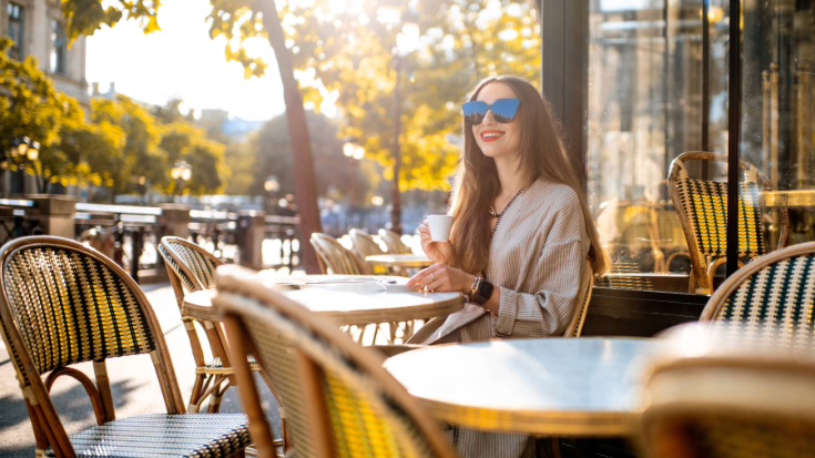 A photograph of a woman alone sitting in the morning sun outside of a Parisian cafe. She has long brown hair down to her chest, is wearing dark sunglasses, is holding a small coffee and is smiling and looking relaxed and happy. She might be a traveller planning her day over a coffee. To illustrate a blog post entitled 'Which Country Has the Best Coffee Culture?'