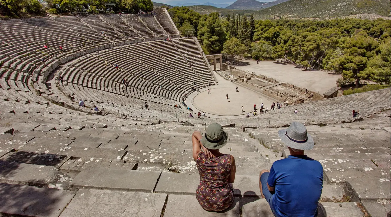 Casal sentado nas arquibancadas do Teatro de Epidauro, sítio arqueológico do século IV a.C. no Peloponeso, Grécia, com a plateia circular e a floresta ao fundo.