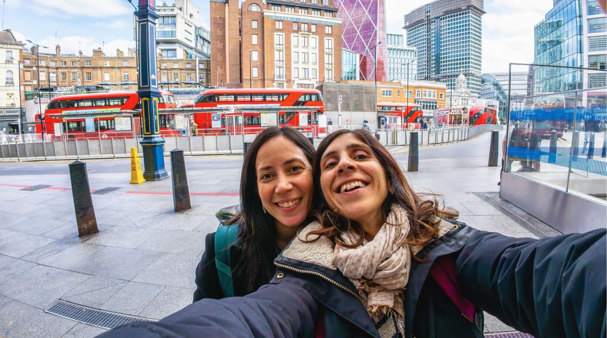 Dos viajeras se hacen una selfie en una calle de ciudad con autobuses rojos y edificios modernos detrás.