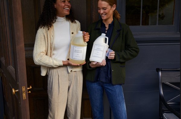 Two people smile and hold The Laundress Signature Detergent Classic Gallon, Fabric Conditioner Classic Gallon, and Measuring Cup.