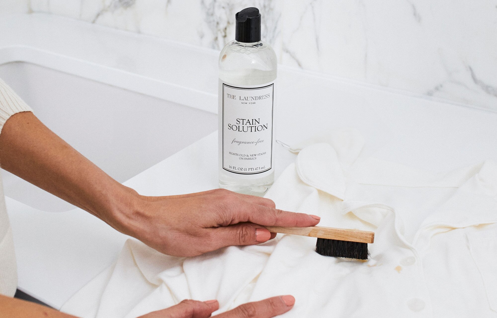 The Laundress Stain Solution stands on a white surface next to hands scrubbing a white shirt with a brush; the background features a marble wall.